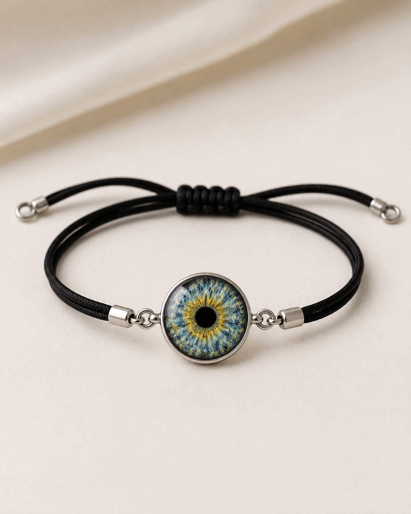 A black cord bracelet holding a silver-set glass iris charm, resting on cream silk